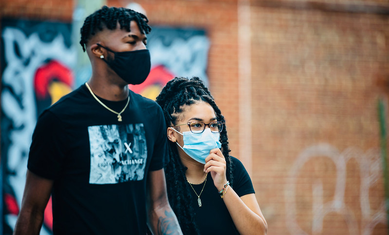 The image features two young individuals standing close to each other, both wearing face masks, suggesting the photo was taken during a time when health precautions were emphasized. The person in the foreground is a woman with glasses and her hair in long, braided locks. She is holding her mask to her chin with one hand, seemingly in contemplation or mid-conversation. The man behind her has a short, curly hairstyle and is wearing a black T-shirt with a graphic and text on it. They are standing in front of a wall with vibrant graffiti, adding a dynamic and colorful backdrop to the scene. The focus on the subjects against the urban art suggests a moment captured during a community event or a casual outdoor gathering.
