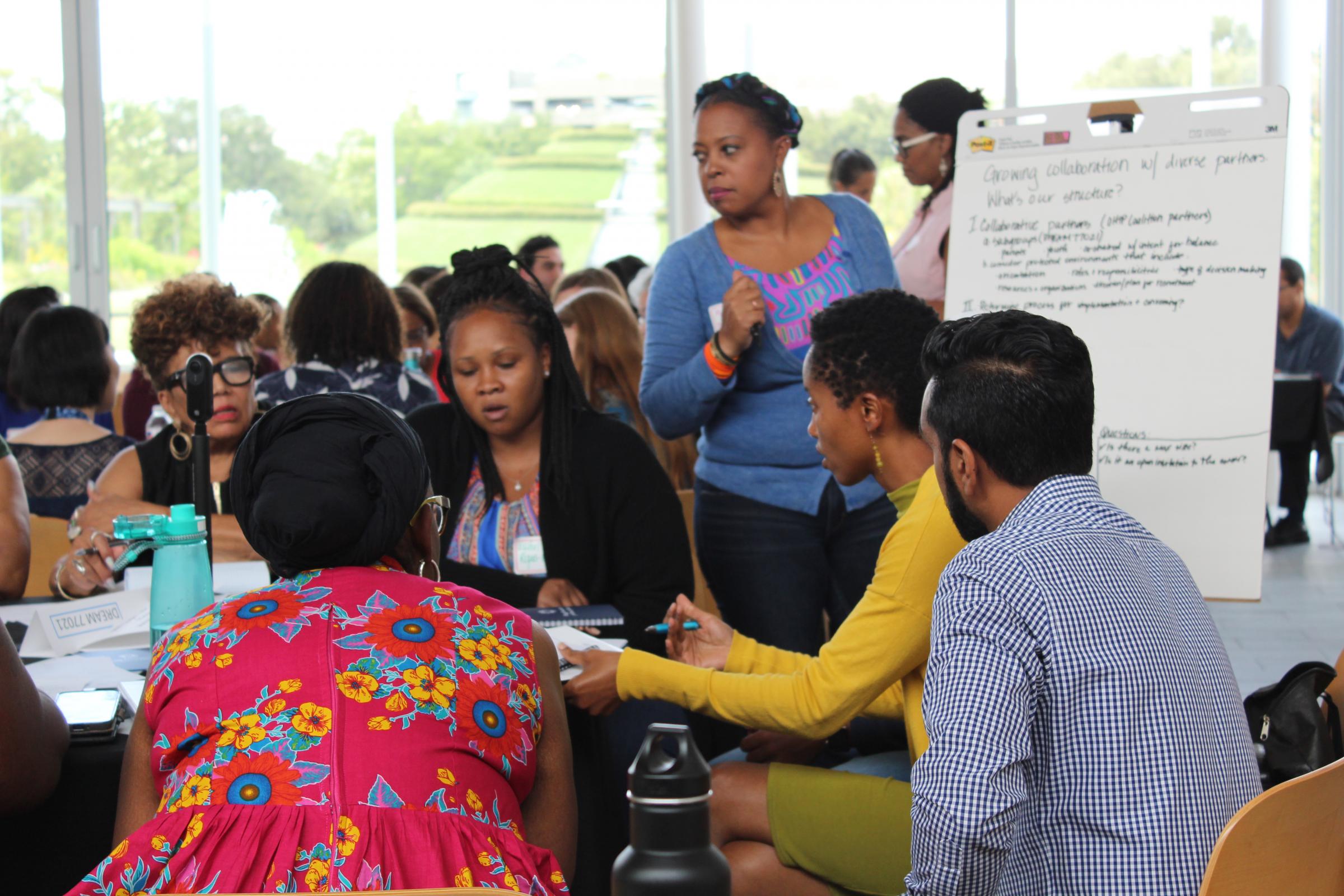 The image captures a diverse group of professionals in a collaborative meeting setting. The foreground shows a woman in a vibrant floral dress, her back to the camera, actively participating in the discussion. Around her, others are seated in groups, some in conversation, while one woman stands, gesturing as if making a point. There's a flip chart in the background with text that reads "Growing collaboration w/ diverse partners," indicating the focus of the meeting. The room is bright and airy with natural light flooding in from large windows, and the participants appear engaged and focused on the task at hand. The setting suggests a workshop or a planning session, with attendees from various backgrounds contributing to a community-oriented project.