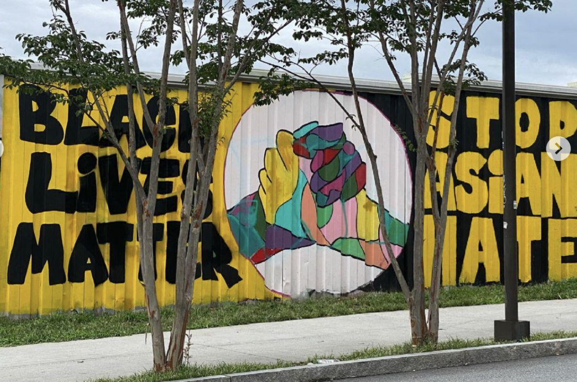 The image depicts a large mural with social justice themes, painted on a corrugated metal surface. On the left, the words "BLACK LIVES MATTER" are painted in bold, black letters on a yellow background. To the right, a continuation of the message reads "STOP ASIAN HATE," with the same black lettering on a yellow background. In the center of the mural, there's a vibrant, multicolored image of two fists clasped together in solidarity. The background for the fists is white, which makes the colorful design stand out. The mural is partially obscured by young trees in the foreground, suggesting it's located in an urban or community setting. The sky is overcast, indicating cloudy weather conditions.