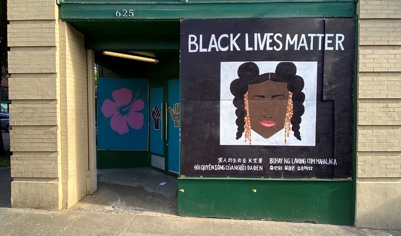 The image shows a mural on the exterior wall of a building with the message "BLACK LIVES MATTER" prominently displayed at the top in large white letters. Below the text is an illustration of a woman with dark skin and stylized hair in buns, with beads hanging from her braids. To the left of the mural is a teal-colored door, and to the left of the door is a smaller mural of a pink flower. On the right side, next to the woman's portrait, there are two hand symbols with one in white and the other in teal, both on a dark background. Beneath the central image, there are phrases written in different languages, including Vietnamese, Tagalog, and Korean, translating to "Black lives matter," demonstrating multicultural support for the movement. The building has a cream-colored brick exterior, and the ground shows a concrete sidewalk.