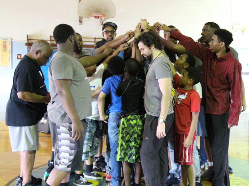 The image shows a diverse group of people, both adults and children, gathered in a gym. They are participating in a team-building exercise or a group huddle, with everyone joining hands together in the air in the center of the circle. The expressions on their faces range from focused to smiling, indicating a positive and collaborative atmosphere. The setting and the activity suggest this could be part of a sports event, a community gathering, or a youth program aimed at fostering teamwork and unity. The participants are casually dressed, appropriate for a physical or social activity in an indoor sports setting.