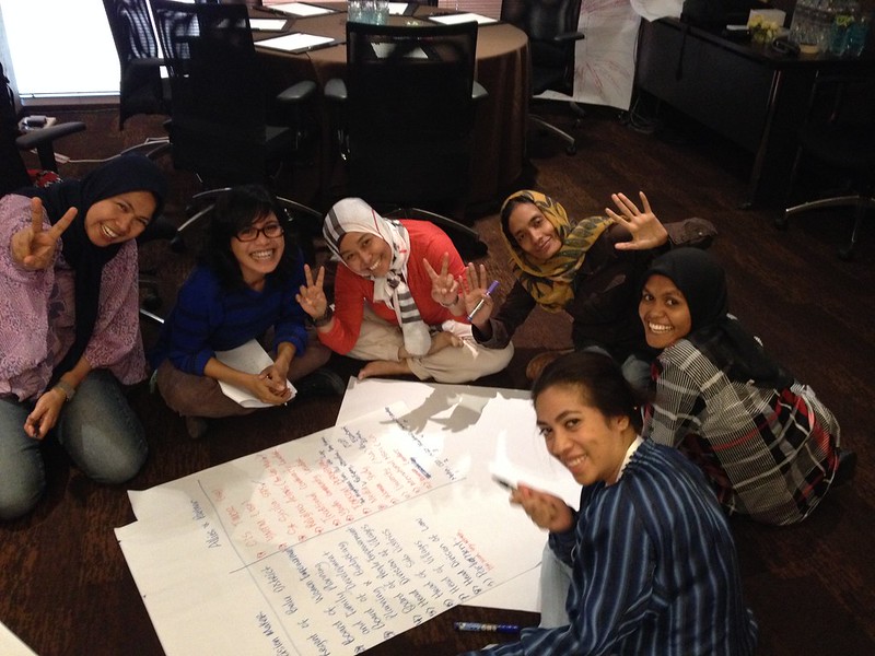 The image shows a group of women sitting on the floor around a large sheet of paper with written notes. They are in a casual office setting, smiling and gesturing peace signs to the camera, indicating a positive, collaborative atmosphere. The women are dressed in comfortable, casual clothes, some wearing hijabs, reflecting cultural diversity. Papers and pens suggest they are brainstorming or involved in a workshop or planning session. The relaxed posture of the women and their cheerful expressions convey a sense of teamwork and camaraderie.