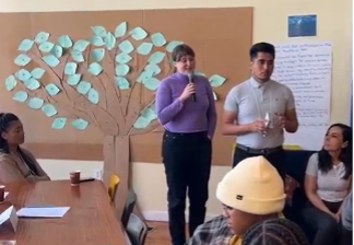 A group of people gathered in a room for a meeting or workshop. Two individuals stand at the front speaking, one holding a microphone and the other holding a small object. Behind them, a large paper tree with green paper leaves is displayed on the wall. Several people sit around a wooden table, listening attentively. There is a large sheet of paper with writing on the wall to the right.