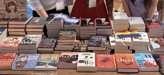 A table is filled with neatly stacked books on display, including titles like Gender Queer, Homegoing, The Color Purple, and How to Burn a Rainbow. Pride flags, including a rainbow and transgender flag, are placed among the books.