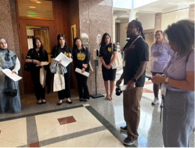 A group of people stands in a hallway with polished floors and marble walls, listening attentively to a man speaking.