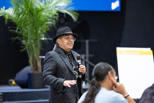 A man wearing a dark hat and suit jacket speaks into a microphone during a presentation. He is smiling and gesturing with his hand, engaging with the audience. Behind him, a large potted plant and a blank flip chart are visible, and another person is seated in the foreground, listening attentively.