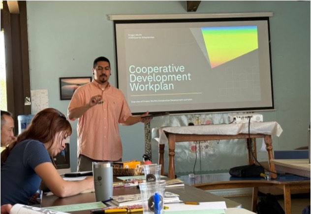 A man stands at the front of a room presenting to a small group, gesturing as a slide titled “Cooperative Development Workplan” is projected on a screen behind him. Several participants sit at tables in the foreground with notebooks and materials, listening and taking notes during the workshop.