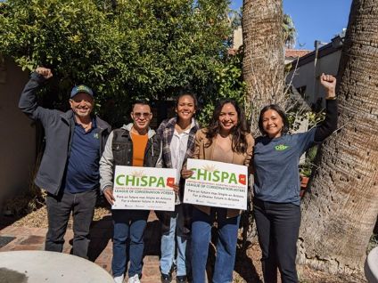 Five people standing outside in a park, two holding signs that say "CHISPA, AZ: Igniting the movement, advancing climate justice; league of conservation voters; for a cleaner future in Arizona"