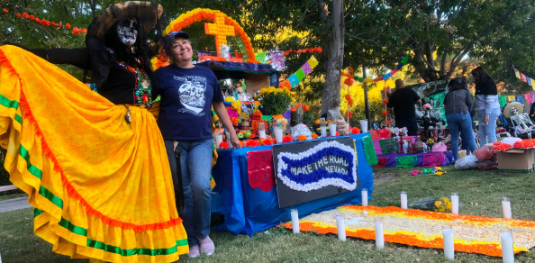 Two people standing in a park next to a table with a blue cloth and various objects to celebrate Día de los Muertos. One person is in denim jeans and a blue shirt. The other person wears a yellow skirt with orange and green stripes at the bottom, a black shirt, and traditional makeup for Día de los Muertos.