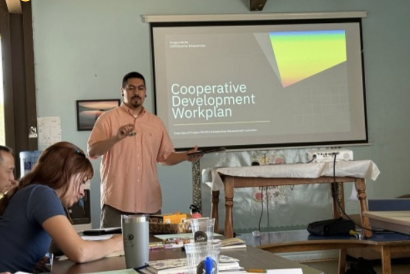 Man presents Cooperative Development Workplan slide to small group in meeting room while participants take notes at table.