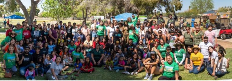 Image of partners gathered on grass from Chispa Arizona; The image shows a large group of people gathered outdoors for what appears to be a community event or social gathering in a park setting. The group is diverse in terms of age and gender, with both children and adults present, many of whom are smiling and posing for the photo. Some individuals are standing while others are seated on the grass. The atmosphere seems casual and cheerful, with some attendees wearing brightly colored clothing and others holding up peace signs, which suggests a friendly and relaxed environment. The presence of banners or signs with text implies that this gathering might be associated with a specific cause or organization, but the details of the signs are not clear from the image. It's a scene that captures a sense of community, engagement, and togetherness.