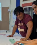 Three community members collaborate at a table reviewing materials, with a presentation slide visible in the background