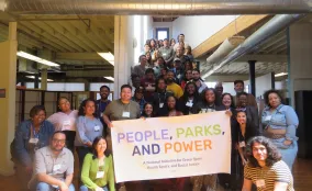Large group of smiling people gathered on a staircase in a bright loft space, holding a "People, Parks, and Power" banner.