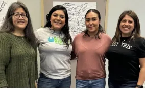 Four women stand together smiling at the camera during a group gathering, with handwritten posters on the wall behind them.