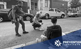 Outreach workers speak with a young person seated on a city sidewalk during a street-based community health outreach visit.