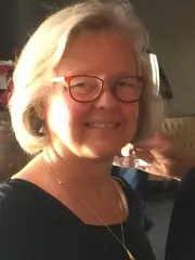 Smiling woman with short gray hair and red glasses standing in a warmly lit interior against a soft background