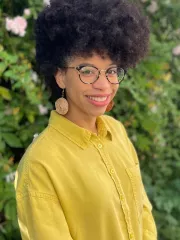 A person with medium-brown skin and a large, rounded curly afro stands outdoors in front of dense green bushes with pink flowers. They wear round glasses, large light-colored circular earrings, and a bright yellow long-sleeve button-down shirt. They are smiling gently and looking toward the camera with their body angled slightly to the side.