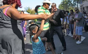 People of different ages dance together outdoors on a sunny day, holding hands in a circle.