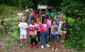 A group of children and adults stand together on a dirt path surrounded by trees and greenery, smiling at the camera during an outdoor community activity.