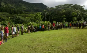 A large group of people stands together on a grassy field with trees and hills in the background.