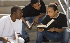 Three adults sit closely together, reading from a book and talking.