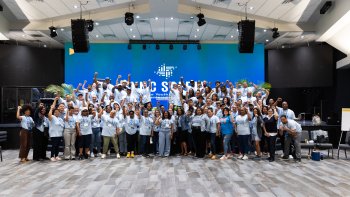 Large group of conference attendees in matching blue shirts pose with raised fists on a stage beneath a large blue screen.