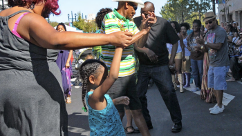 People of different ages dance together outdoors on a sunny day, holding hands in a circle.