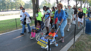 A group of community members, including adults and children, stand together outdoors holding handmade signs during a public gathering. The group appears to be participating in a community action or advocacy event in a park-like setting.