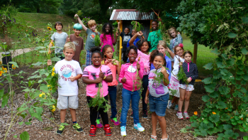 A group of children and adults stand together on a dirt path surrounded by trees and greenery, smiling at the camera during an outdoor community activity.
