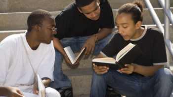 Three adults sit closely together, reading from a book and talking.