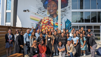 Group of diverse adults gathered outdoors, smiling in front of a large mural of a Black woman at a community center.