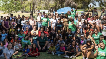 The image shows a large group of people gathered outdoors for what appears to be a community event or social gathering in a park setting. The group is diverse in terms of age and gender, with both children and adults present, many of whom are smiling and posing for the photo. Some individuals are standing while others are seated on the grass. 