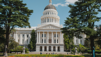 California State Capitol building in Sacramento framed by tall trees, with landscaped gardens and a bright blue sky.