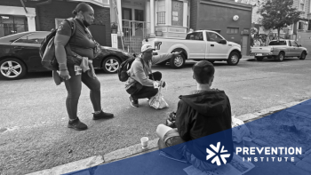 Outreach workers speak with a young person seated on a city sidewalk during a street-based community health outreach visit.