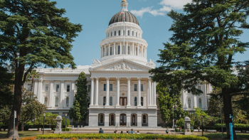 Front view of California State Capitol in Sacramento, white dome and columns framed by trees, people on lawn, blue sky.