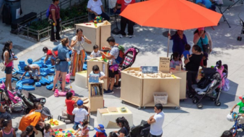 Families and children gather around play tables and activity stations at an outdoor community plaza event.