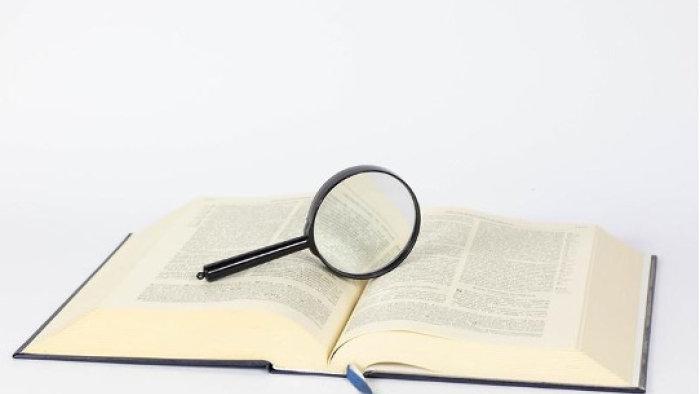 The image shows a large, open hardcover book resting on a flat surface with pages that appear to contain a lot of text, suggesting it may be a reference book or encyclopedia. On top of the open pages lies a round magnifying glass with a black handle, which is used to enlarge text for easier reading. The background is plain and white, emphasizing the book and magnifying glass. The overall scene conveys a theme of research, study, or the pursuit of knowledge.