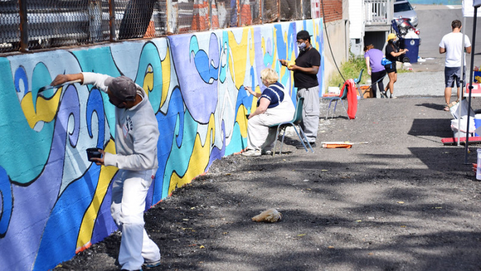 The image shows a group of people engaged in a community mural painting activity. They are outdoors, working on a wall that already features a partially completed abstract design with blue and yellow elements. Some individuals are standing while painting, and others are seated. The painters are wearing casual clothing, and some have protective clothing like overalls. The atmosphere appears to be casual, with some people focused on their painting and at least one person checking a phone. 