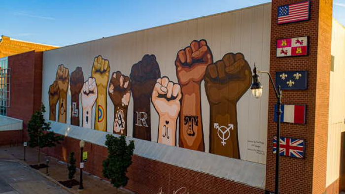 The image shows a mural of raised fists in different skin tones, each adorned with a letter to spell out "SOLIDARITY." Beside the fists are various flags, including the United States flag, indicating international unity or support for a cause. The mural is on a large building wall, viewed under a clear sky, and suggests themes of diversity, unity, and collective strength.