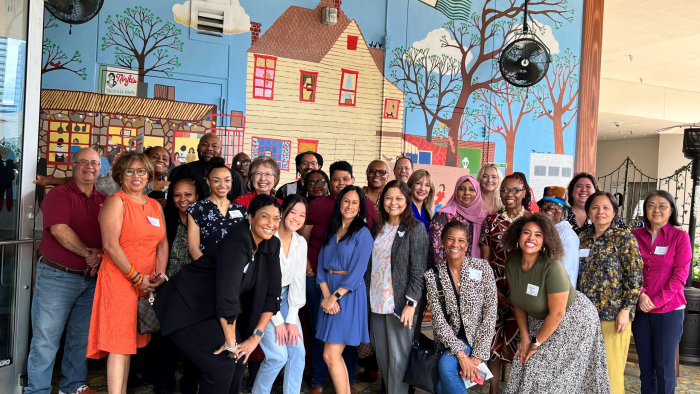 A group of 27 people pose for a photo in front of a mural marked "Ninfa's The Original 1973," suggesting a gathering at a historical or significant location. The diverse group, displaying a mix of ages, races, and attire, seems to be at a social or professional event, indicated by their casual to business-casual clothes and name tags. They stand in a well-lit indoor space that appears to be a restaurant or public venue.
