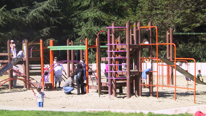 Children play on a colorful playground with slides, ladders, and climbing bars while adults watch nearby in a sandy park.