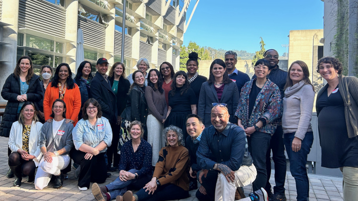 Group of adults posing outdoors in a courtyard, smiling together in front of a modern building under a clear blue sky.