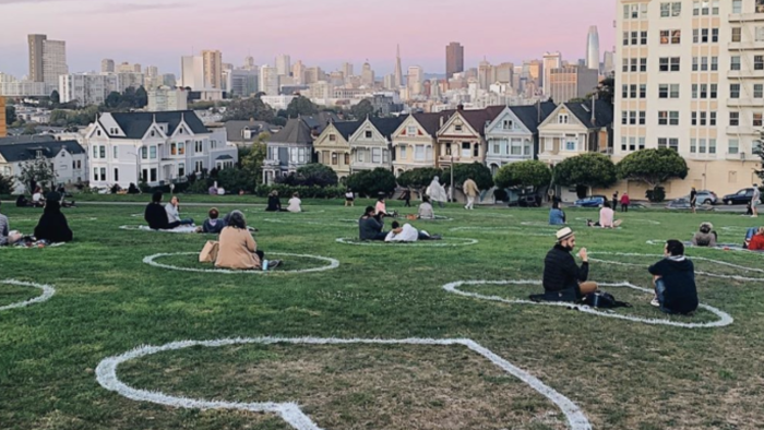 People sit 6 ft apart in chalk hearts on a grassy park lawn with San Francisco’s Painted Ladies and skyline in the background at sunset.