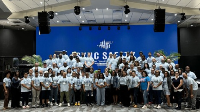 Large group of participants gathered on stage in front of blue SYNC wall, smiling together at a convening event