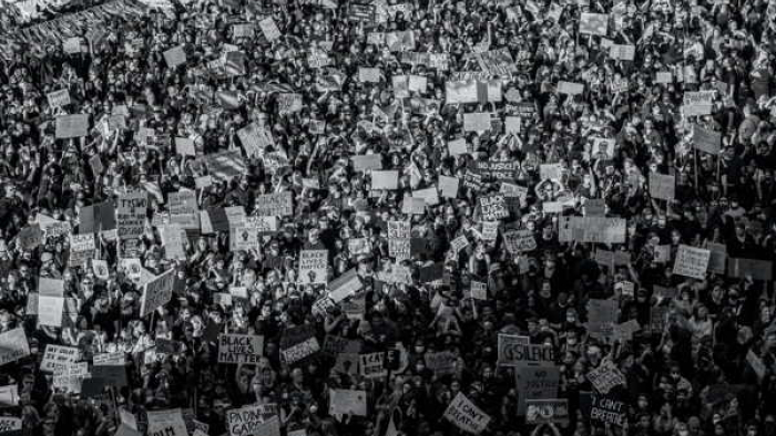 The image is a black and white photograph capturing a dense crowd of protesters. The mass of people is holding up various signs, creating a sea of placards that suggest a large-scale demonstration or public event focused on a cause or multiple issues. The absence of color emphasizes the unity and collective voice of the participants. The photo conveys the magnitude and intensity of public demonstrations and the power of collective action.