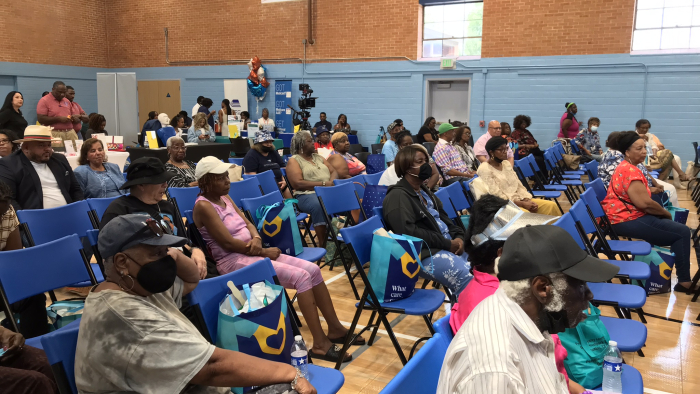 A community event taking place in a gymnasium with rows of seated attendees. Most individuals are older adults, and several are wearing masks. The attendees sit on blue chairs, some holding bags with health-related logos. A table in the back features informational materials, and staff are seen standing in the background. The setting is bright, with natural light coming through large windows.